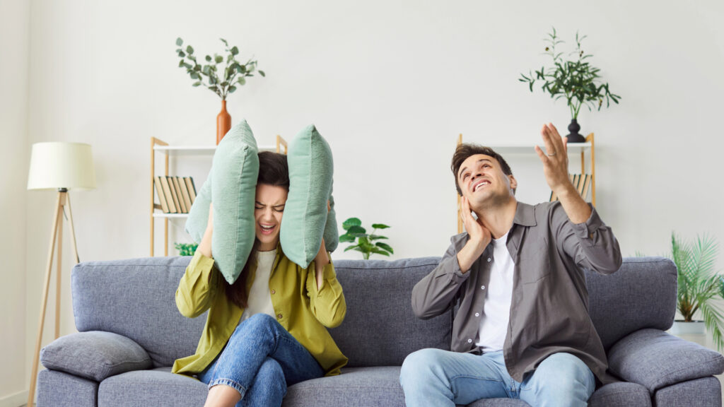 A woman holds pillows over her ears to block out loud neighbors while a man sitting on the couch next to her holds his ears.